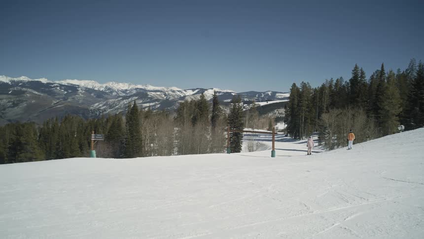 A wide, slow pan reveals two skiers pausing on a snowy slope, with a breathtaking view of a vast mountain range under a clear blue sky