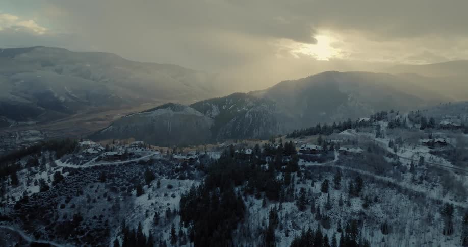 Sunlight breaks through moody clouds over a vast, snow-covered mountain landscape with a small village nestled in the hills