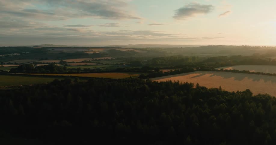 An aerial drone shot flies over the peaceful countryside with rolling hills, forests, and fields bathed in the warm light of a beautiful sunset