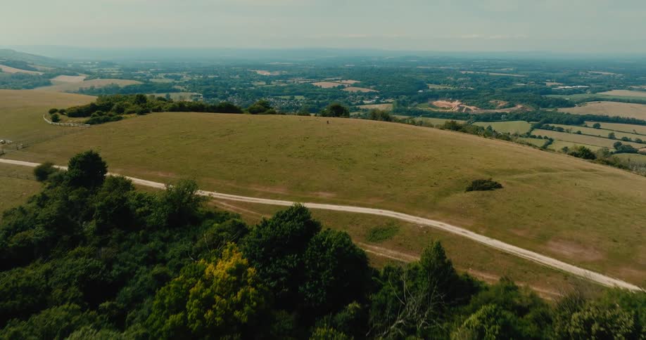 A soaring aerial shot reveals the stunning, vast English countryside after rising over a lush green, tree-covered hill on a beautiful day