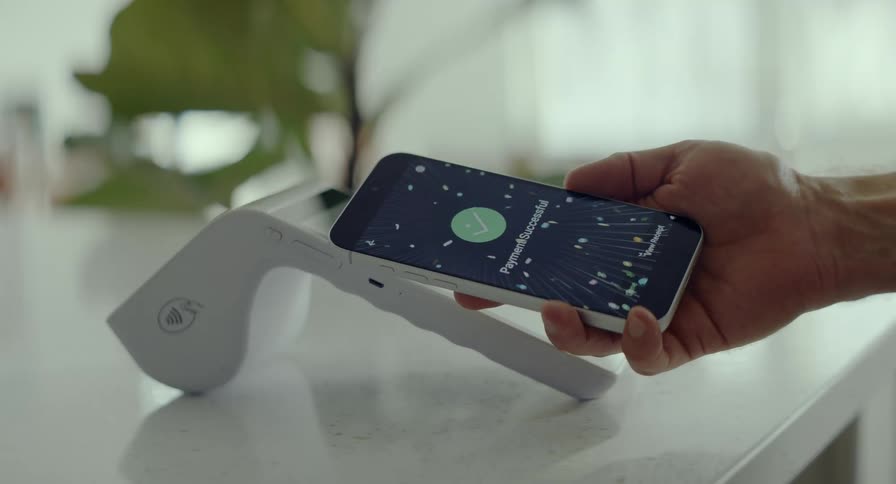 A man's hand holds a smartphone over a contactless payment terminal showing a successful transaction with a celebratory animation