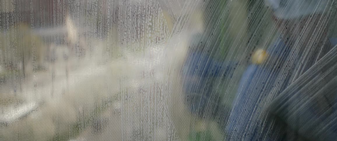 A window washer uses a squeegee to clean soapy suds off a skyscraper's glass window, with another worker reflected in the background