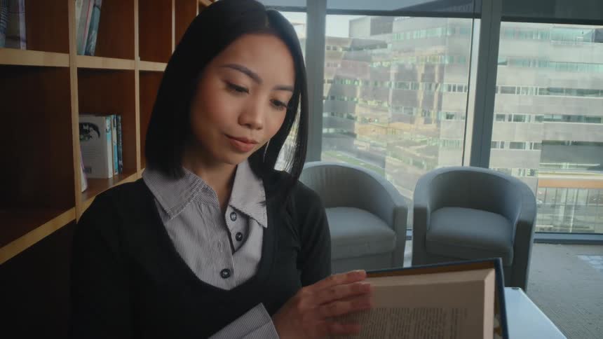 A young Asian woman pulls a book from a bookshelf in a modern library with a large window overlooking the city