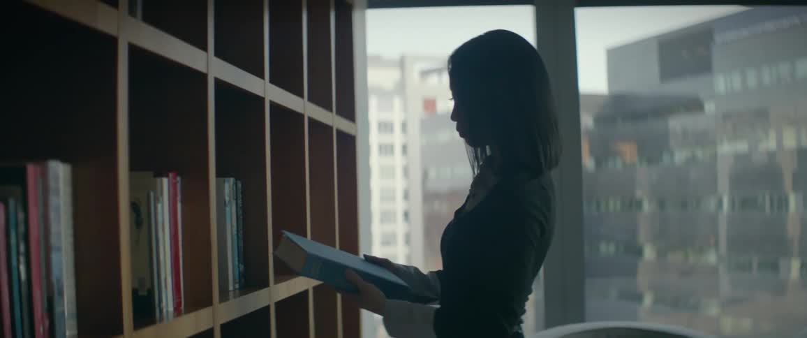 A professional woman in a modern high-rise office selects a book from a wooden bookshelf and opens it to read