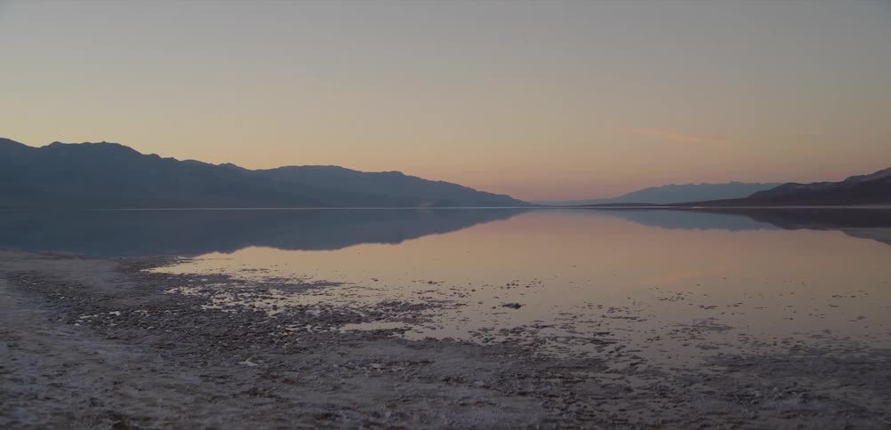 The tranquil waters of a desert lake perfectly mirror the distant mountains under a soft-hued sunrise sky