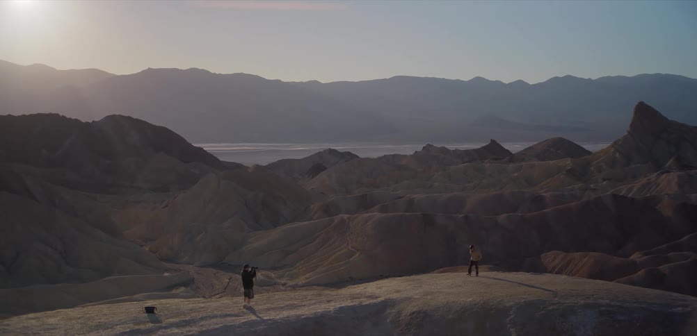 A photographer with a tripod films a person walking along a ridge overlooking the stunning badlands of Death Valley at sunrise