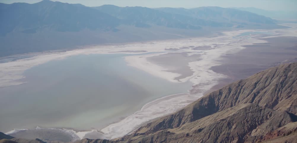 An expansive high-angle view from a rugged mountain slowly reveals a vast desert valley and salt flat lake