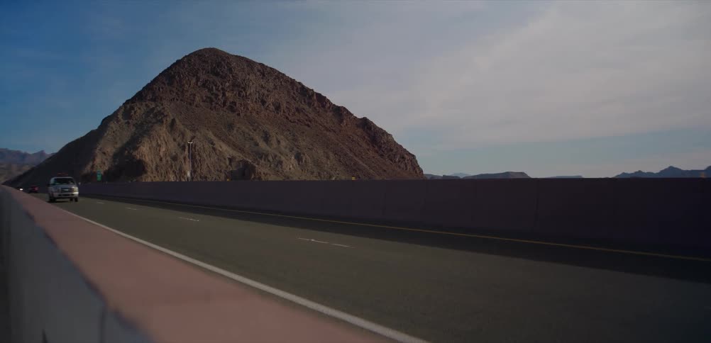 Vehicles, including multiple pickup trucks, drive in slow motion along a desert highway with a large mountain in the background
