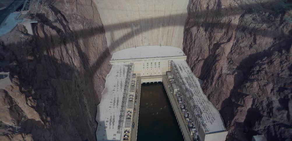 A high-angle static shot looking down at the Hoover Dam powerhouse and the Colorado River, with the shadow of the Memorial Bridge above