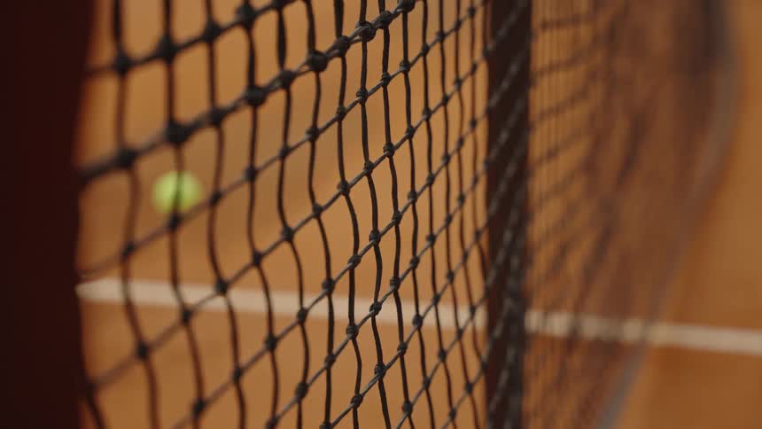 A yellow tennis ball bounces in slow motion behind the net on a red clay tennis court