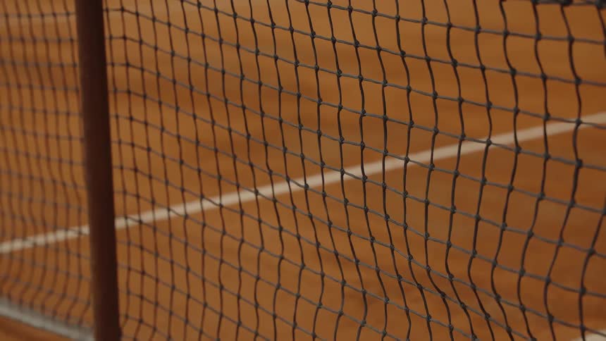 A yellow tennis ball bounces in slow motion on a clay court, seen through the net