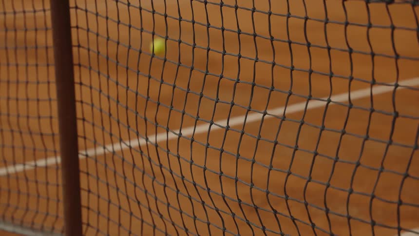 A tennis ball is served and bounces on a clay court in slow motion, viewed through the net