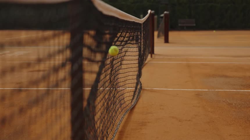 A tennis ball repeatedly bounces off the net on an empty clay court in slow motion