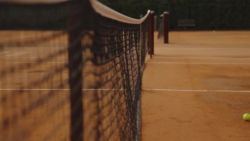 A tennis ball hits the net in slow motion on an empty clay court during a beautiful golden hour