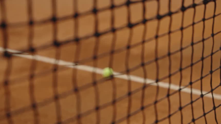 A slow rack focus shot from a tennis net to a single tennis ball on a clay court