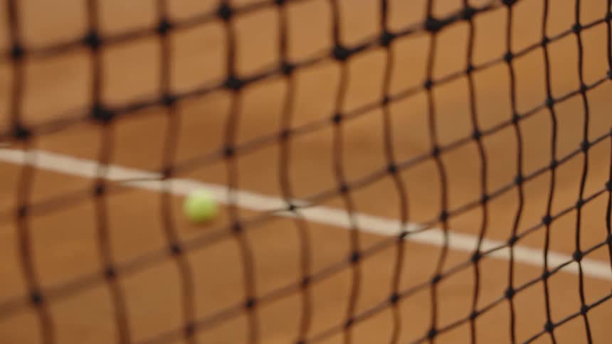 A detailed focus pull from a tennis net to a single yellow tennis ball resting on a clay court