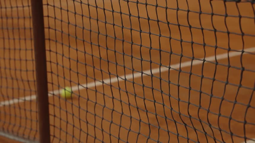 A yellow tennis ball bounces in slow motion on a clay court, seen through the mesh of the net