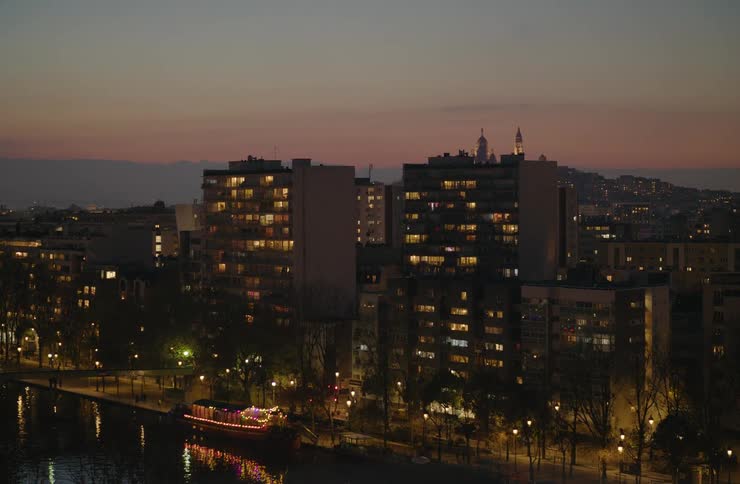 A wide shot of a European city skyline at dusk, with lights twinkling in apartment buildings and a tour boat floating on the river below