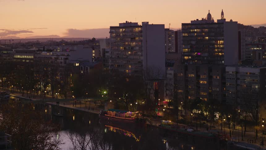 A wide static shot of a calm city canal at sunset, with lights illuminating the surrounding apartment buildings and a tour boat below