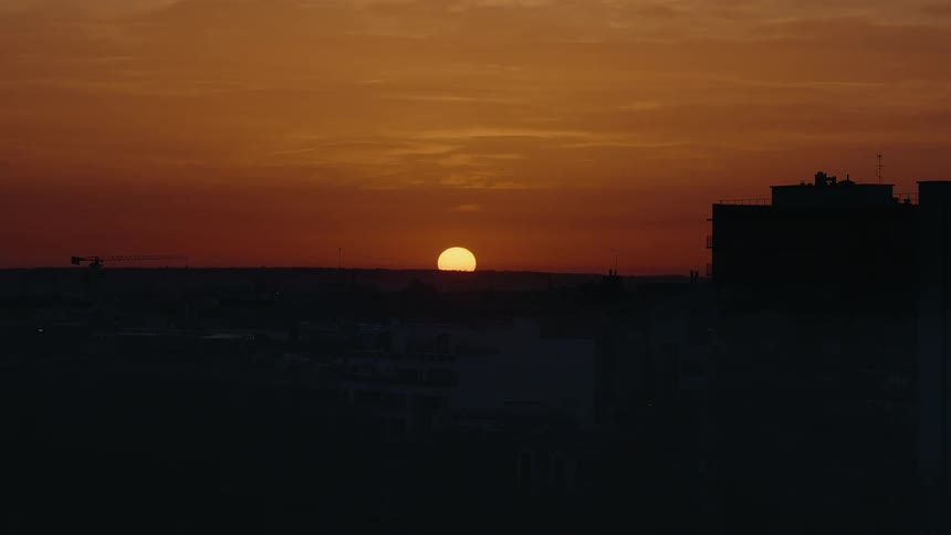 A beautiful time-lapse shows the sun setting behind a silhouetted city skyline against a vibrant orange sky