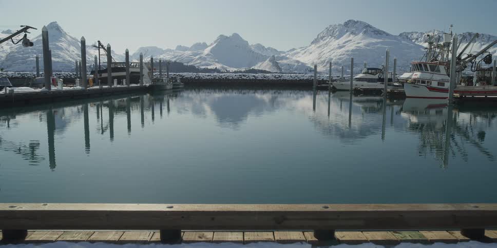 A tranquil winter scene at a boat harbor with snow-covered mountains reflecting perfectly in the calm, glassy water on a bright sunny day