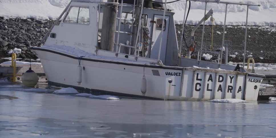 A white fishing boat named Lady Clara from Valdez, Alaska sits docked and frozen in an icy harbor during a cold winter day