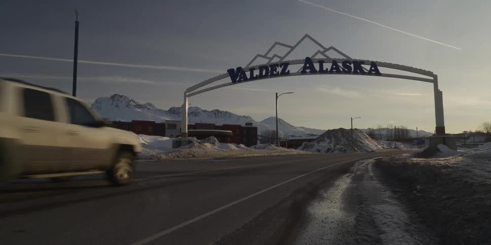 A truck drives into the snowy mountain town of Valdez, Alaska, passing under the iconic welcome sign at sunrise