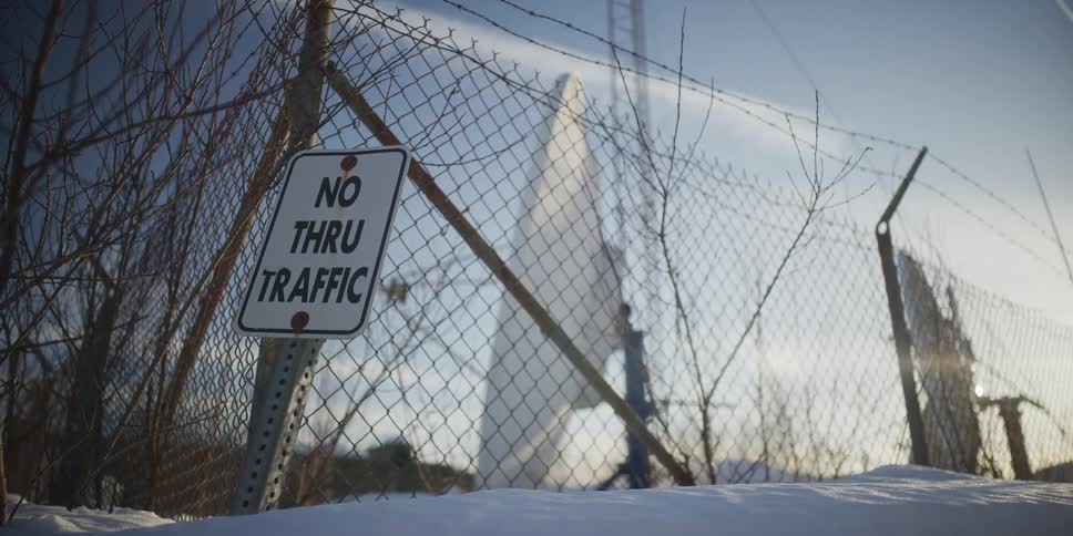 A 'No Thru Traffic' sign on a snowy chain-link fence with a large satellite dish in the background at sunset