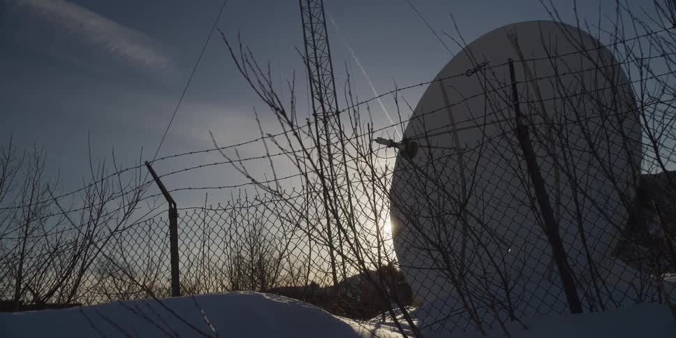 A satellite dish stands behind a barbed wire fence in the snow, silhouetted by the winter sun