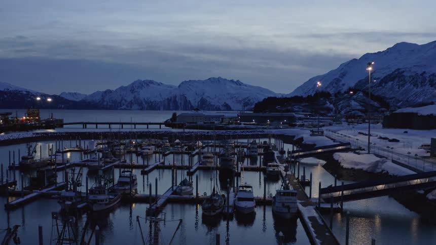 A tranquil aerial pullback reveals a snowy Alaskan harbor filled with docked fishing boats during the cold blue light of twilight