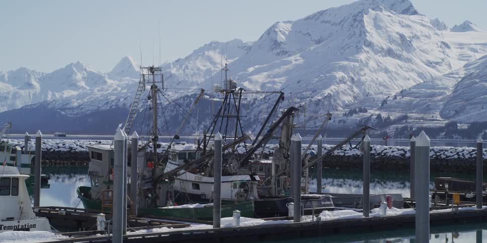A panning shot of snow-covered boats in a tranquil harbor with a dramatic backdrop of towering, snow-capped mountains