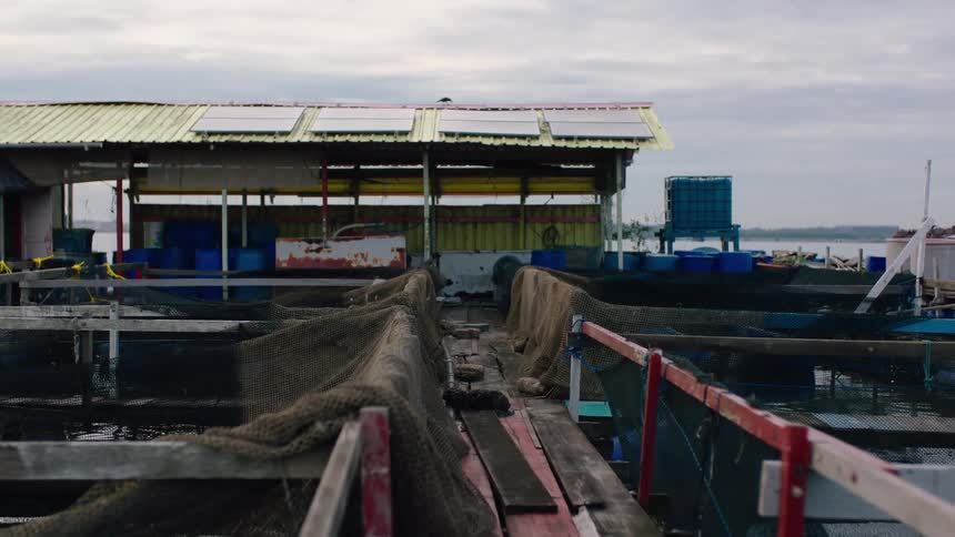 A static view of a rustic floating fish farm with nets and solar panels on an overcast day