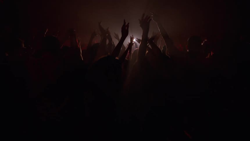 A silhouetted crowd of fans dance with their hands in the air at a dark, smoky concert in slow motion