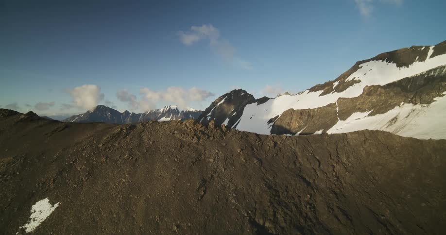 A cinematic aerial shot slowly reveals a vast, remote mountain range with snow-covered peaks under a clear blue sky