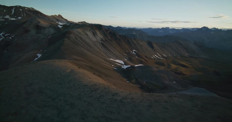 Epic aerial footage pulls back over a vast and rugged mountain range with patches of snow during a golden sunrise
