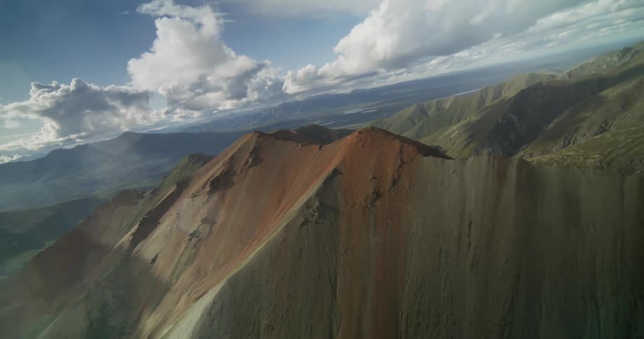 A canted aerial perspective reveals a majestic, rust-colored mountain range beneath a blue and cloudy sky