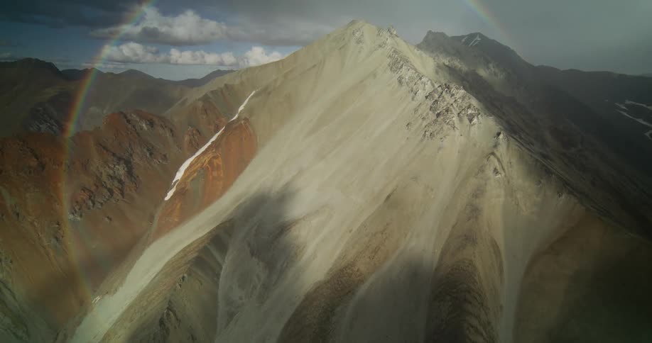 An epic aerial view captures a vibrant double rainbow arching over a majestic, sunlit mountain peak amidst dramatic, stormy clouds