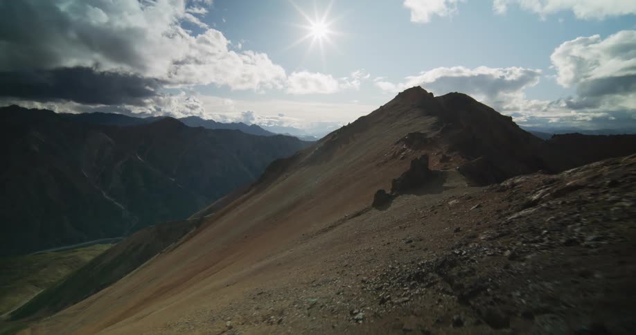 An aerial view flies over a dramatic and rocky mountain ridgeline on a sunny day with clouds in the sky
