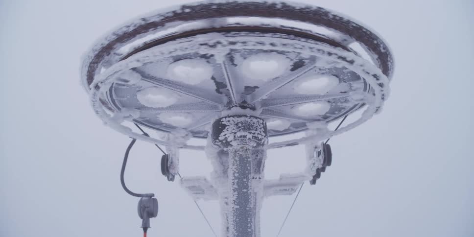 A frozen ski lift pulley wheel covered in frost and snow rotates during a heavy blizzard at a mountain resort