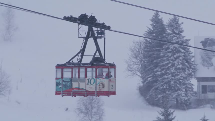A red gondola lift slowly glides through a heavy snowstorm over a snow-covered alpine village