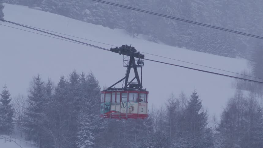 A red cable car moves slowly across a snowy, tree-lined landscape during a snowstorm