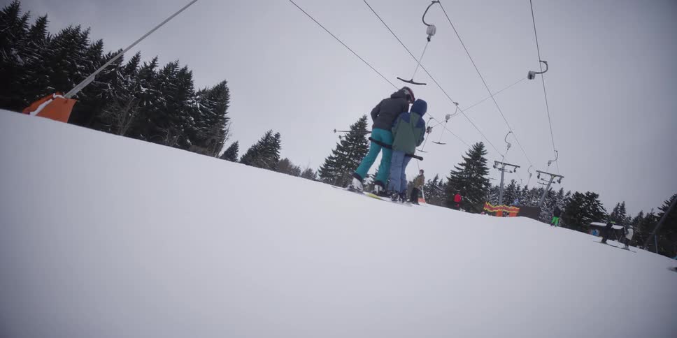 Two skiers ride a T-bar lift up a snow-covered hill at a ski resort on a cloudy winter day