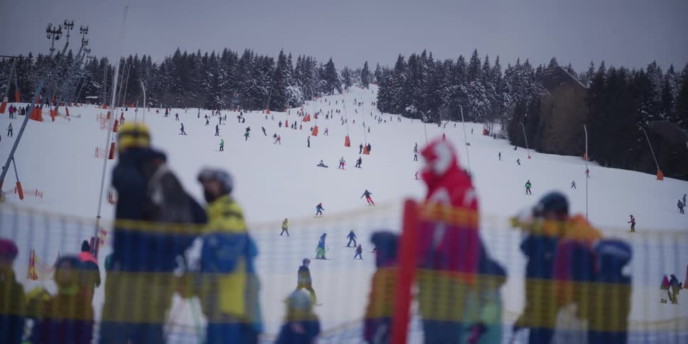 A wide, static view from behind a safety net of a bustling ski slope filled with people enjoying winter sports on an overcast day