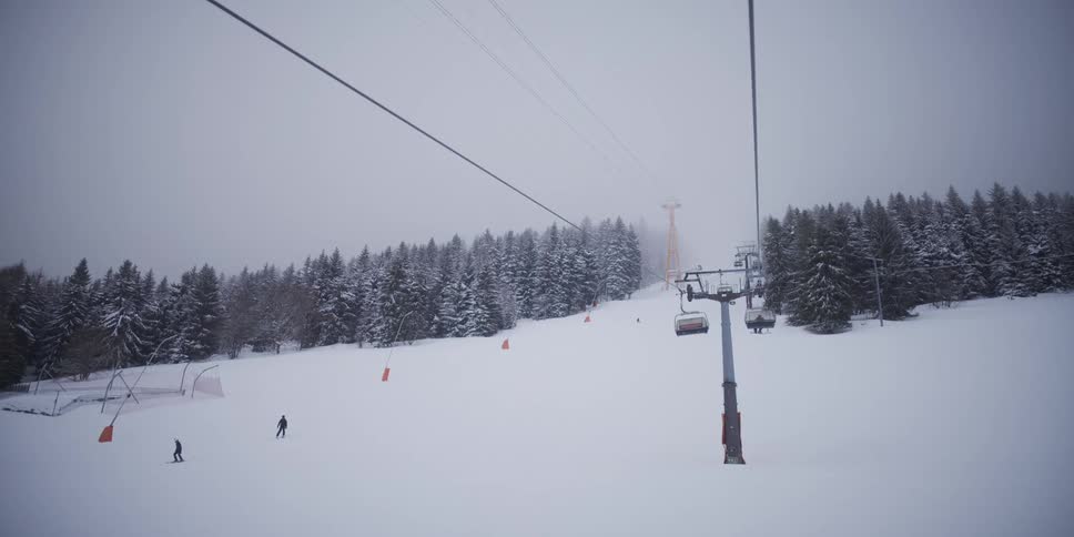 A serene, slow-motion point-of-view shot from a ski lift gliding over a snowy slope with skiers on a foggy winter day
