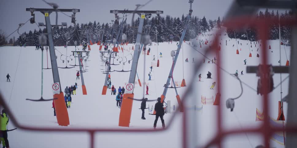 Skiers and snowboarders ride T-bar lifts up a crowded mountain slope at a busy ski resort on an overcast winter day