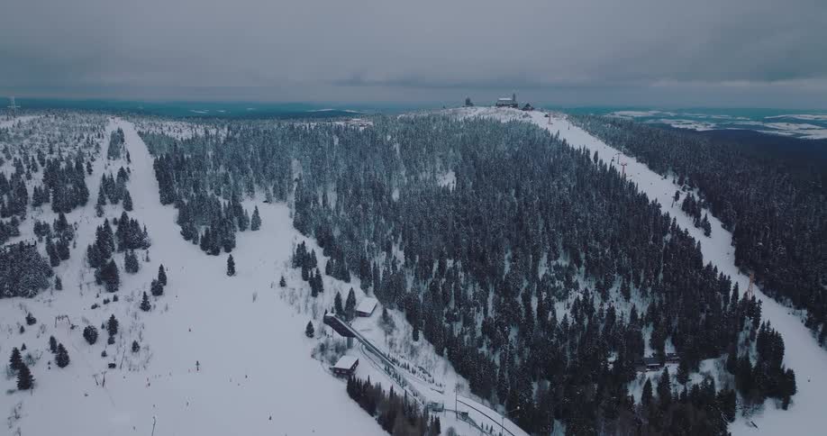 High aerial view slowly reveals a sprawling ski resort on a snow-covered mountain with dense pine forests under a cloudy winter sky