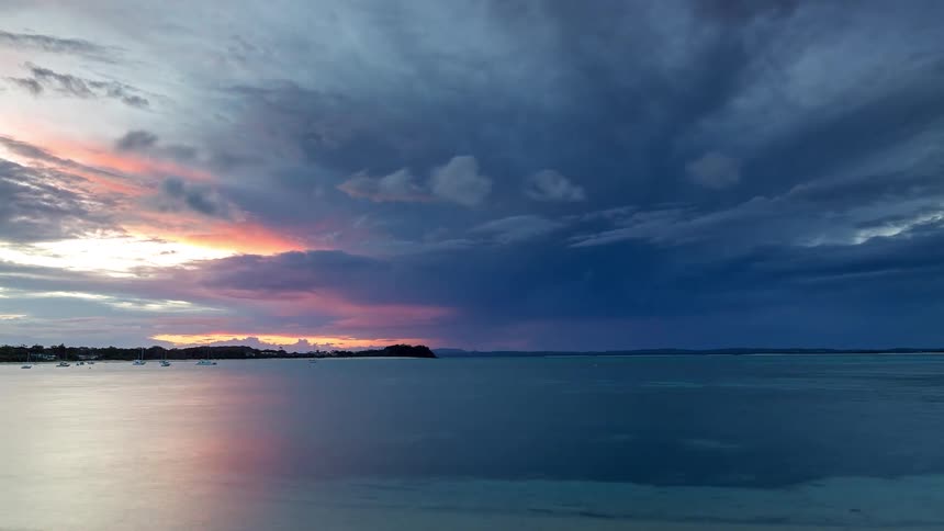 A dramatic timelapse captures a moody sunset with vibrant colors reflecting on the calm ocean as dark clouds roll in