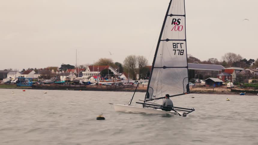 Slow motion side-view of a person sailing a small dinghy along the coastline of a seaside town on a grey, overcast day