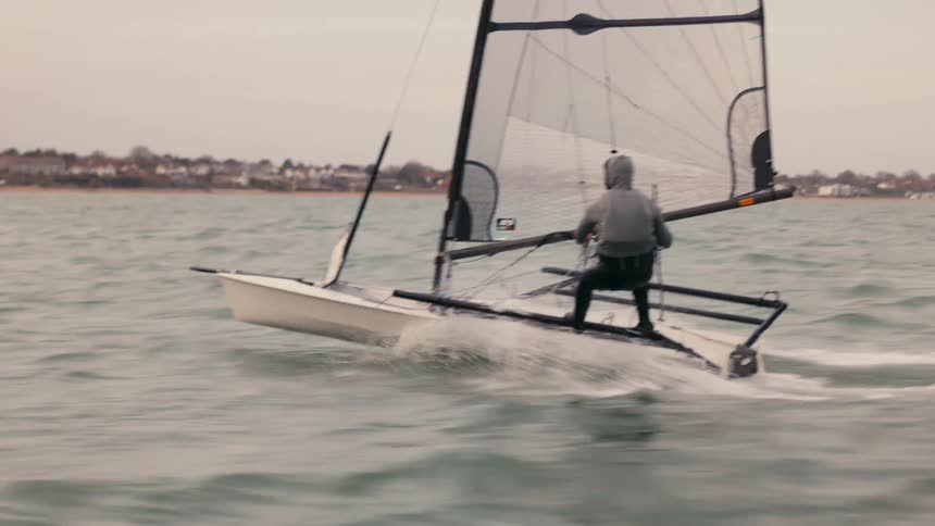 A lone sailor skillfully navigates a high-performance dinghy at speed along the coast on a grey day