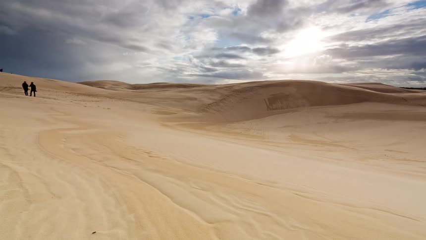 Timelapse of people exploring the vast, windswept sand dunes of a desert under a dramatic and cloudy sky